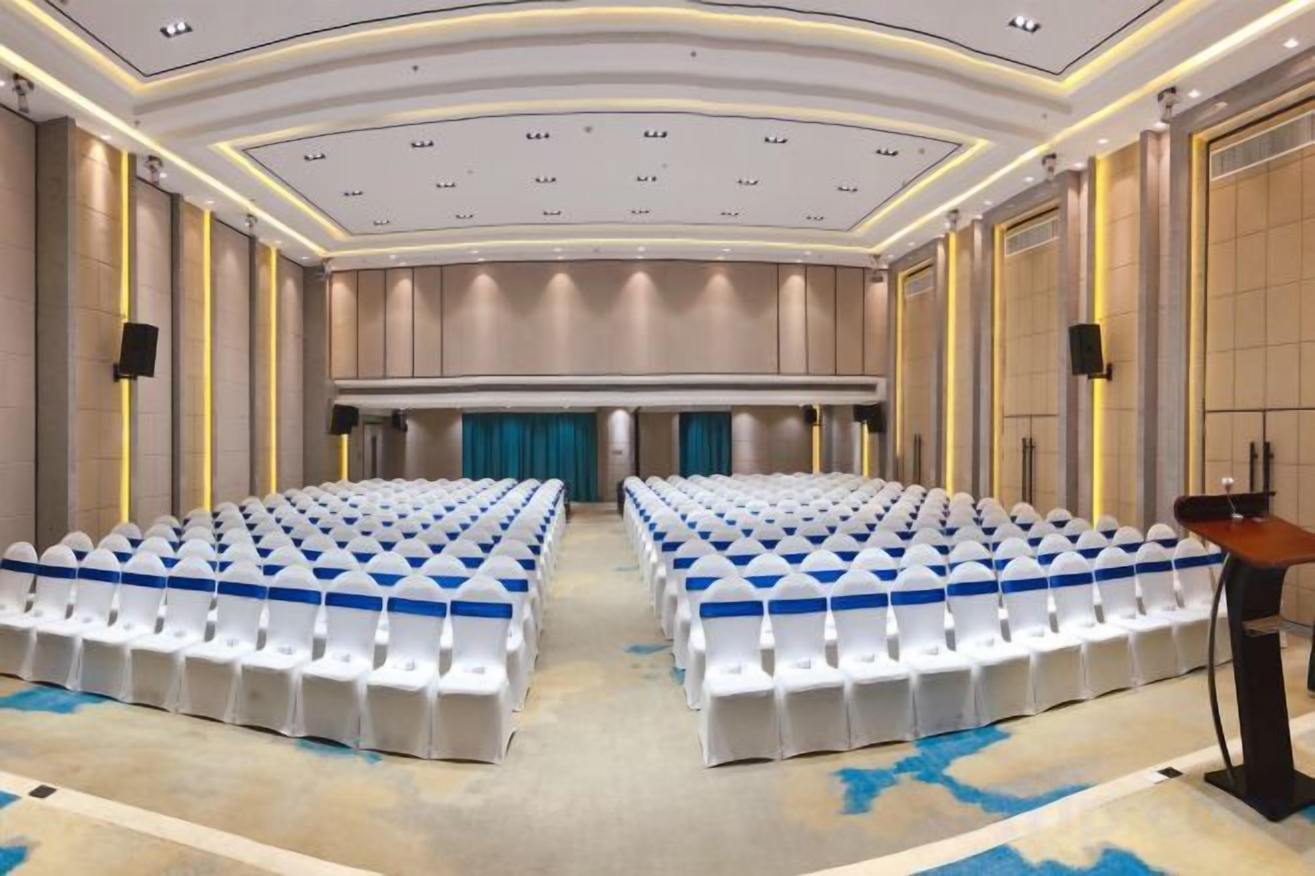 A conference room with rows of white chairs, a podium, and a stage with a blue curtain.