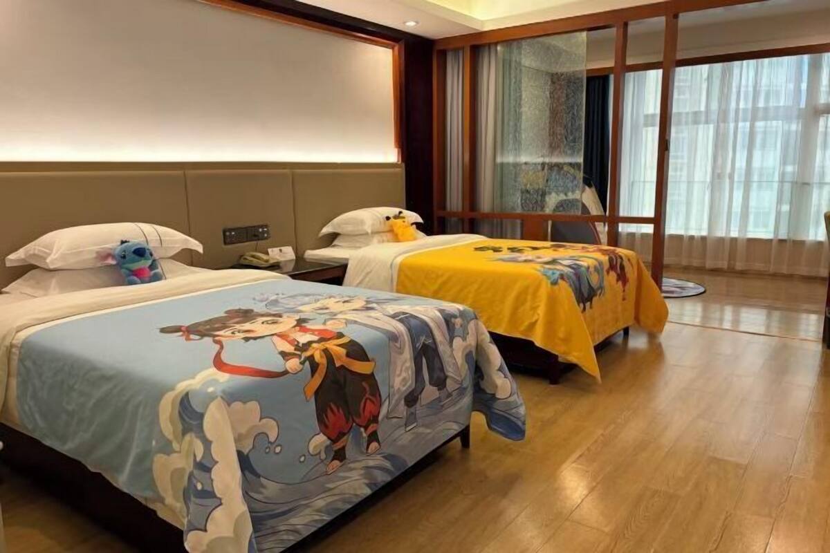 Une chambre d’hôtel avec deux lits, chacun recouvert d’une couvre-lit à motifs de dessins animés différents. La pièce est dotée d’une grande fenêtre avec des rideaux légers, d’un plancher en bois et d’une petite table ronde.