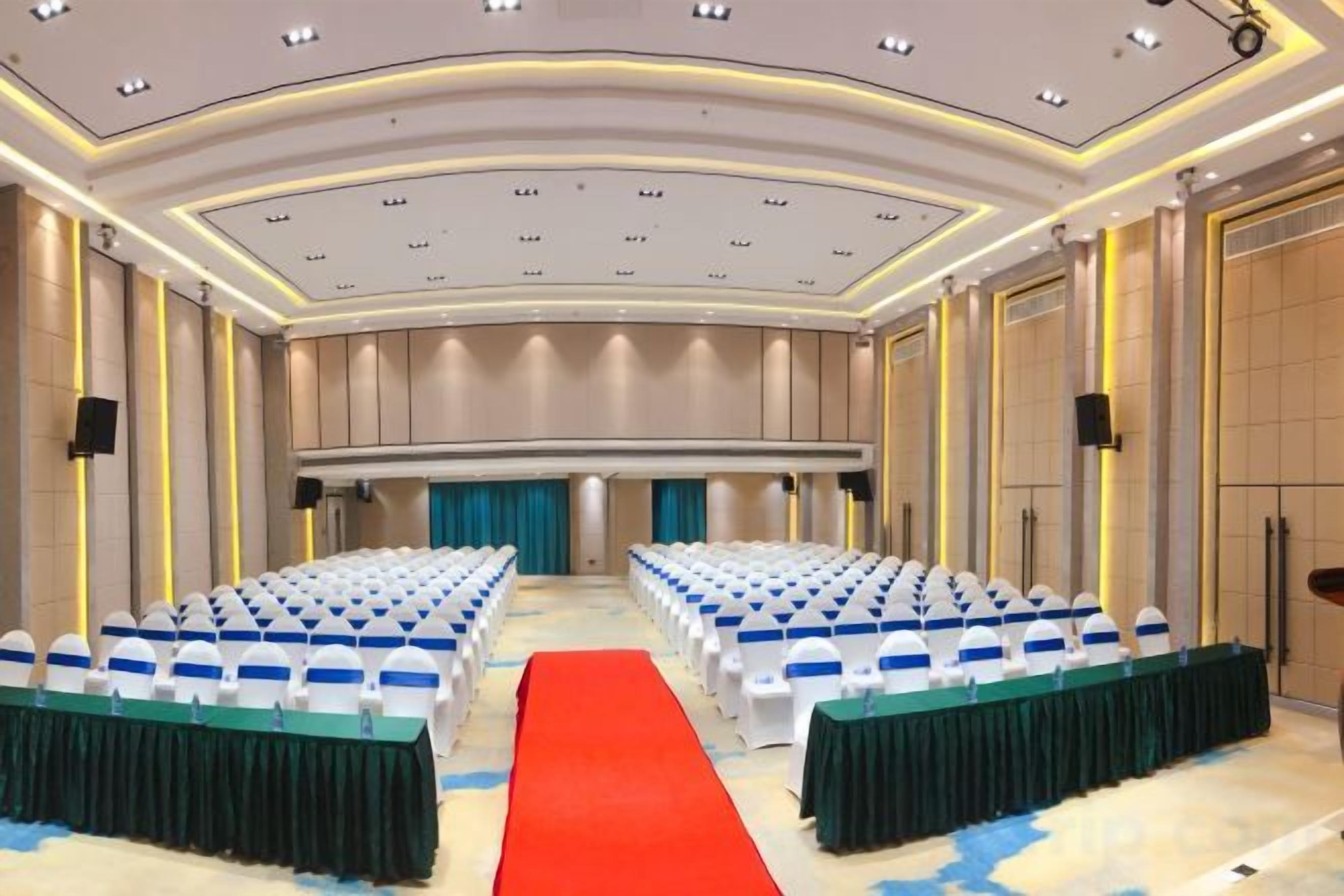 Une salle de conférence avec des rangées de chaises blanches, une moquette rouge et une scène surmontée d’un rideau bleu.