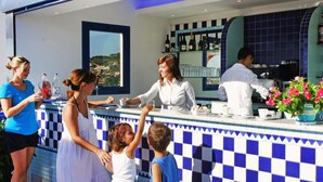 Un coin bar avec un mur recouvert de carreaux bleus et blancs formant un motif à carreaux, une vue sur le paysage par une fenêtre et un comptoir où une personne sert des boissons.