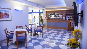 Un hall d’hôtel avec un sol à carreaux, un coin bar et un espace repas avec des chaises bleues et une table blanche.