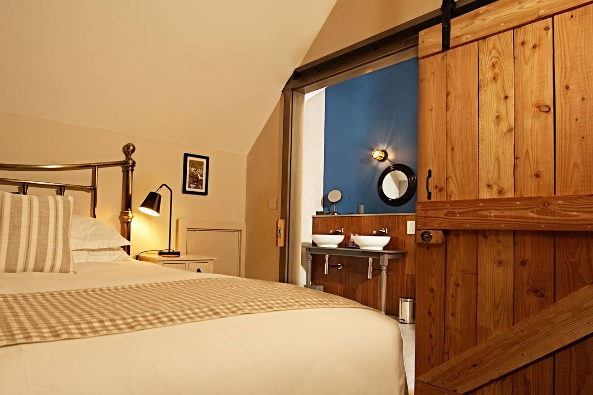 A bedroom with a bed, bedside lamp, wooden wardrobe, and a bathroom with a sink and mirror.