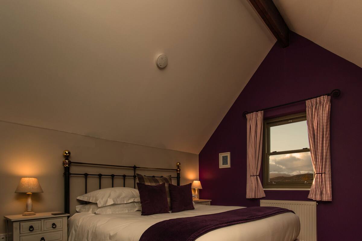 A bedroom with a bed, two bedside tables with lamps, a window with checkered curtains, and a view of the outdoors.