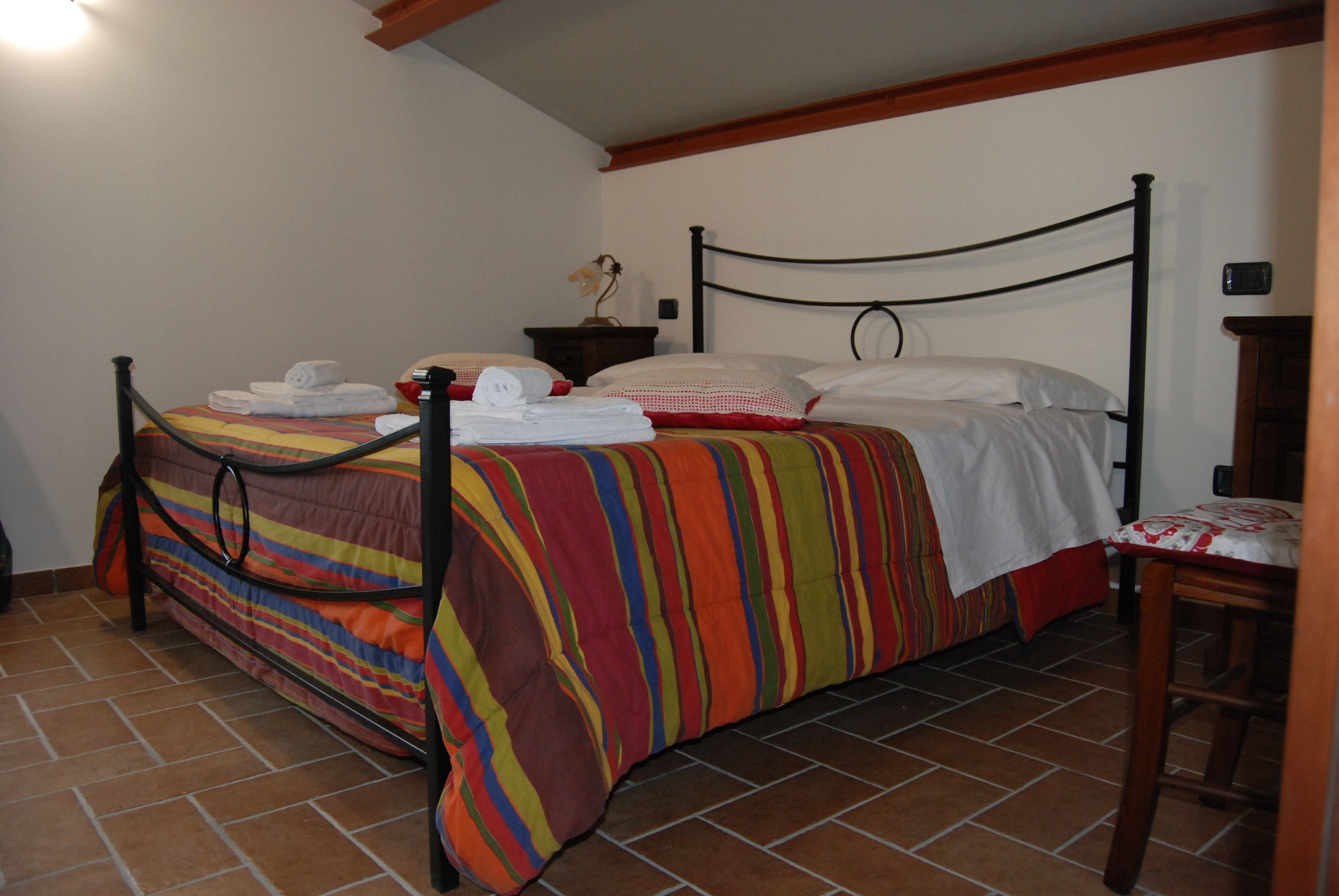 A single bed with a colorful striped bedspread, a small bedside table with a lamp, and a chair with a patterned cushion.