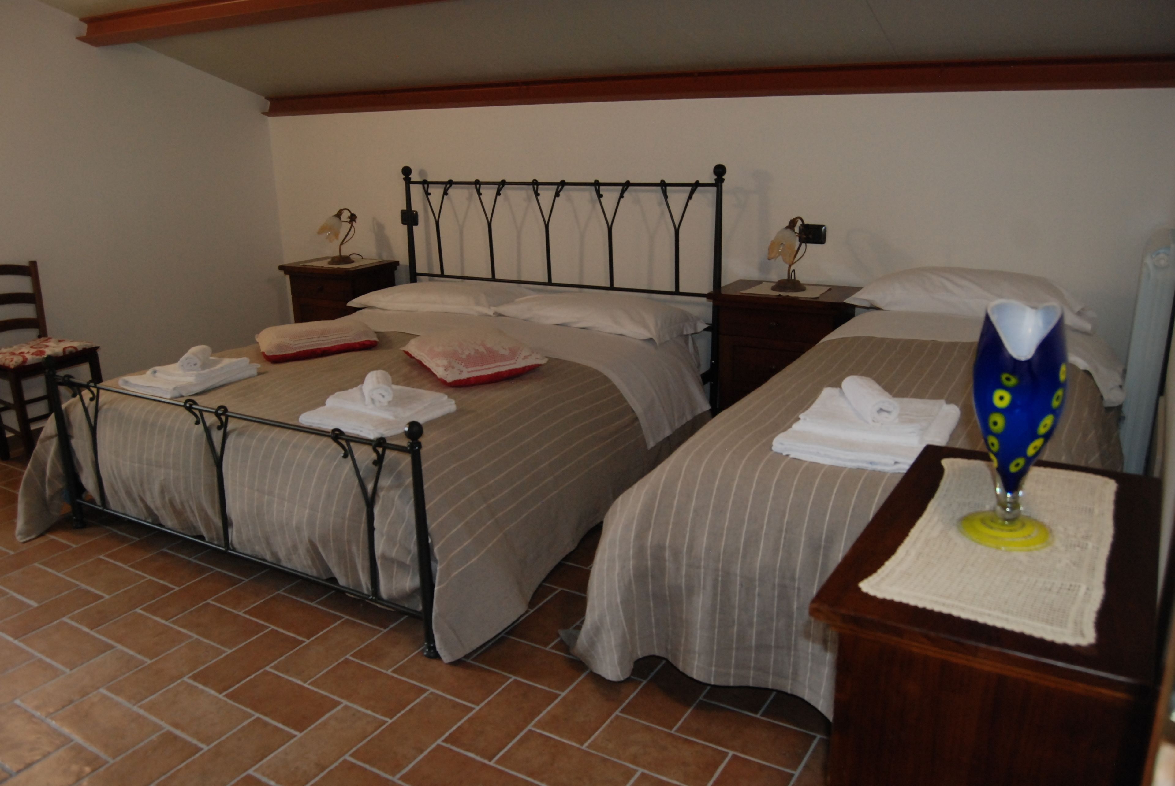 A hotel room with two beds, each with a metal frame and striped bedding. There are bedside tables with lamps and a small wooden nightstand.