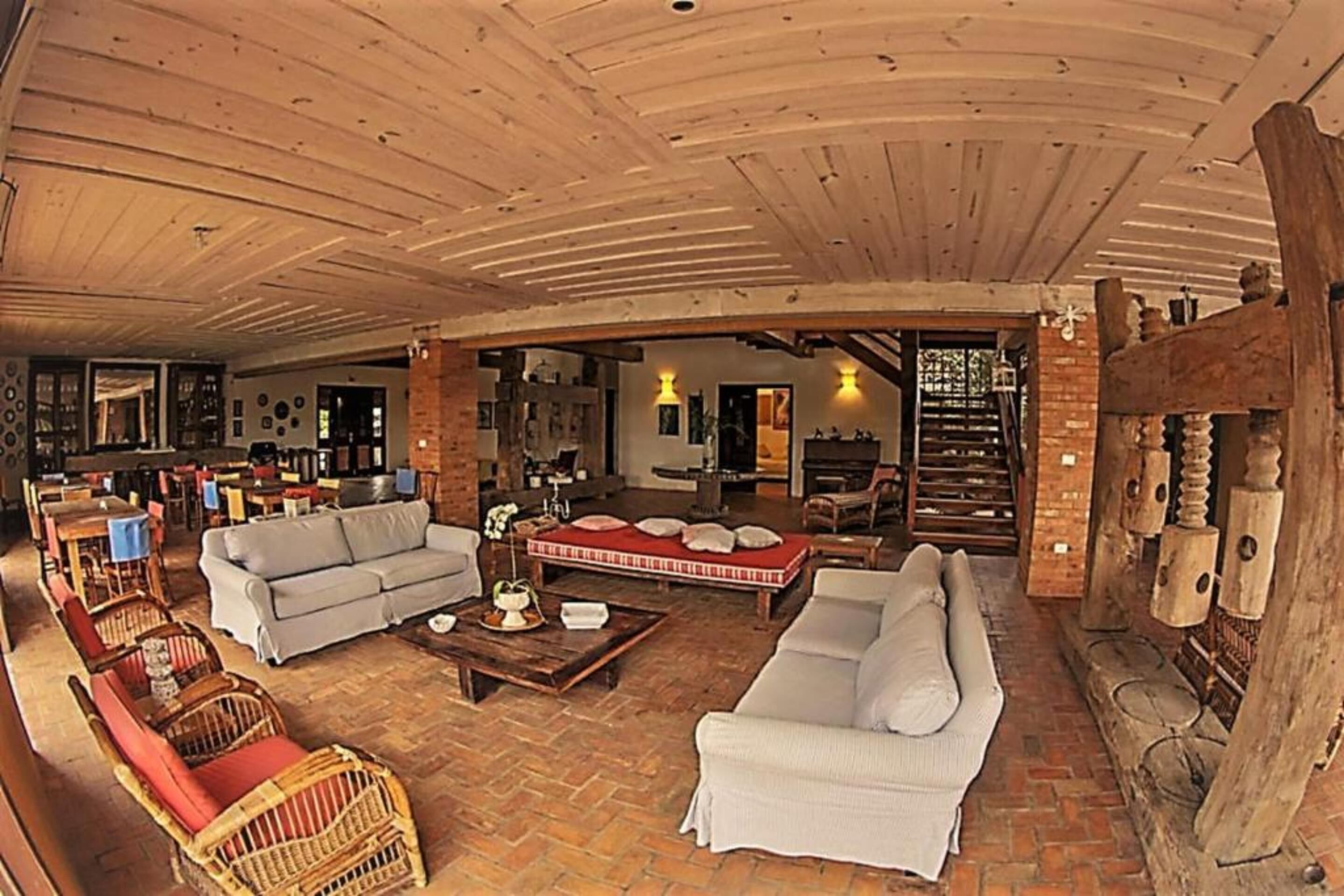 A spacious indoor area with a high wooden ceiling, furnished with sofas and wicker chairs, and a brick wall with a wooden beam structure.