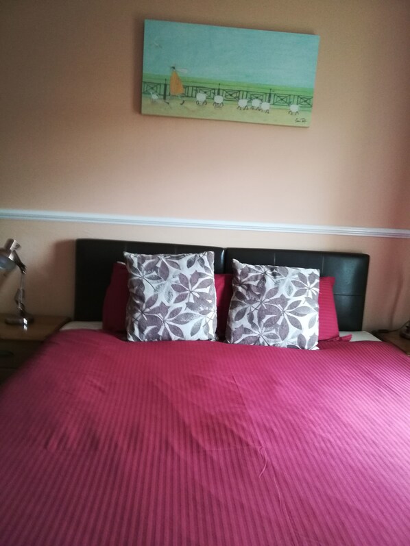 A bed with a red bedspread and two patterned pillows, a nightstand with a lamp, and a painting on the wall. - Bryn Mor Beach Hotel (Caernarfon)