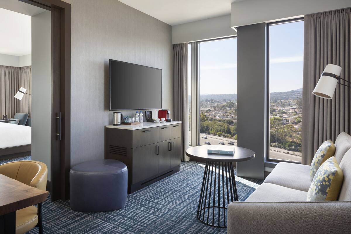 Habitación de hotel con cama, escritorio, sofá, televisor, una mesita y un amplio ventanal con vistas a la ciudad.