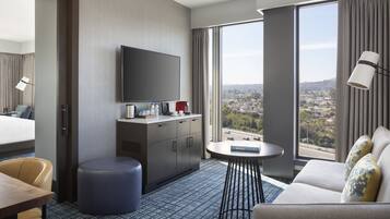 A hotel room with a bed, a desk, a sofa, a TV, a small table, and a large window offering a city view.