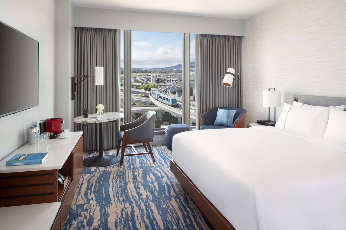 Habitación de hotel con una cama grande, un escritorio con silla, una mesita y vistas a una autopista y al paisaje urbano a través de amplios ventanales.