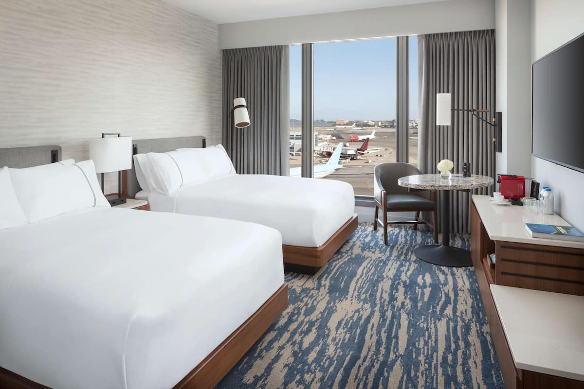 Habitación de hotel con dos camas, un escritorio, una silla, un televisor y vista al aeropuerto.