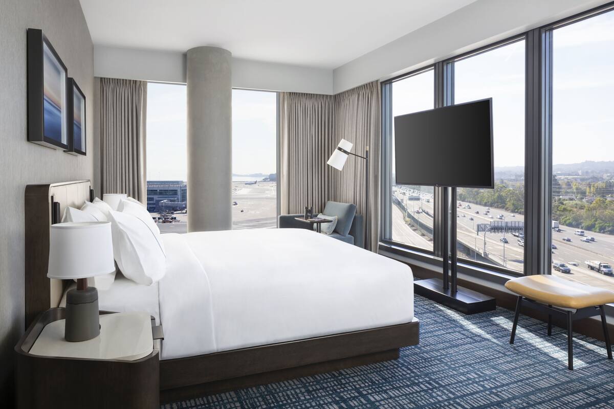 Habitación de hotel moderna con una cama grande, un escritorio, una silla, un televisor y vistas de un paisaje urbano a través de amplias ventanas.
