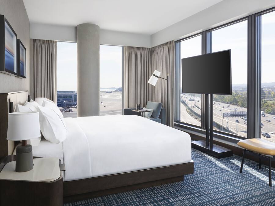 A modern hotel room with a large bed, a desk, a chair, a TV, and a view of a cityscape through large windows.