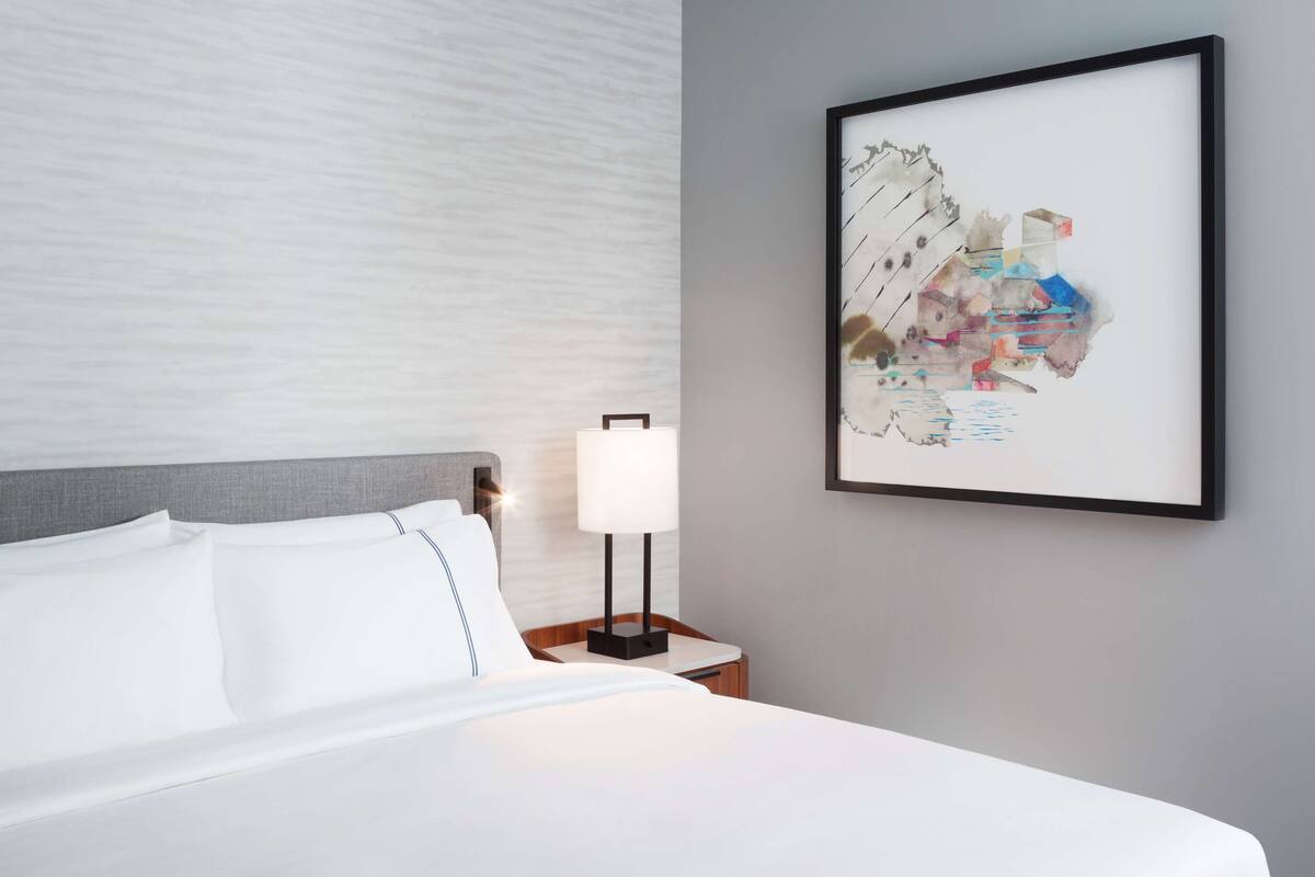 Un cuarto de hotel con una cama bien hecha, una obra de arte abstracta enmarcada en la pared y una mesita de noche con una lámpara.
