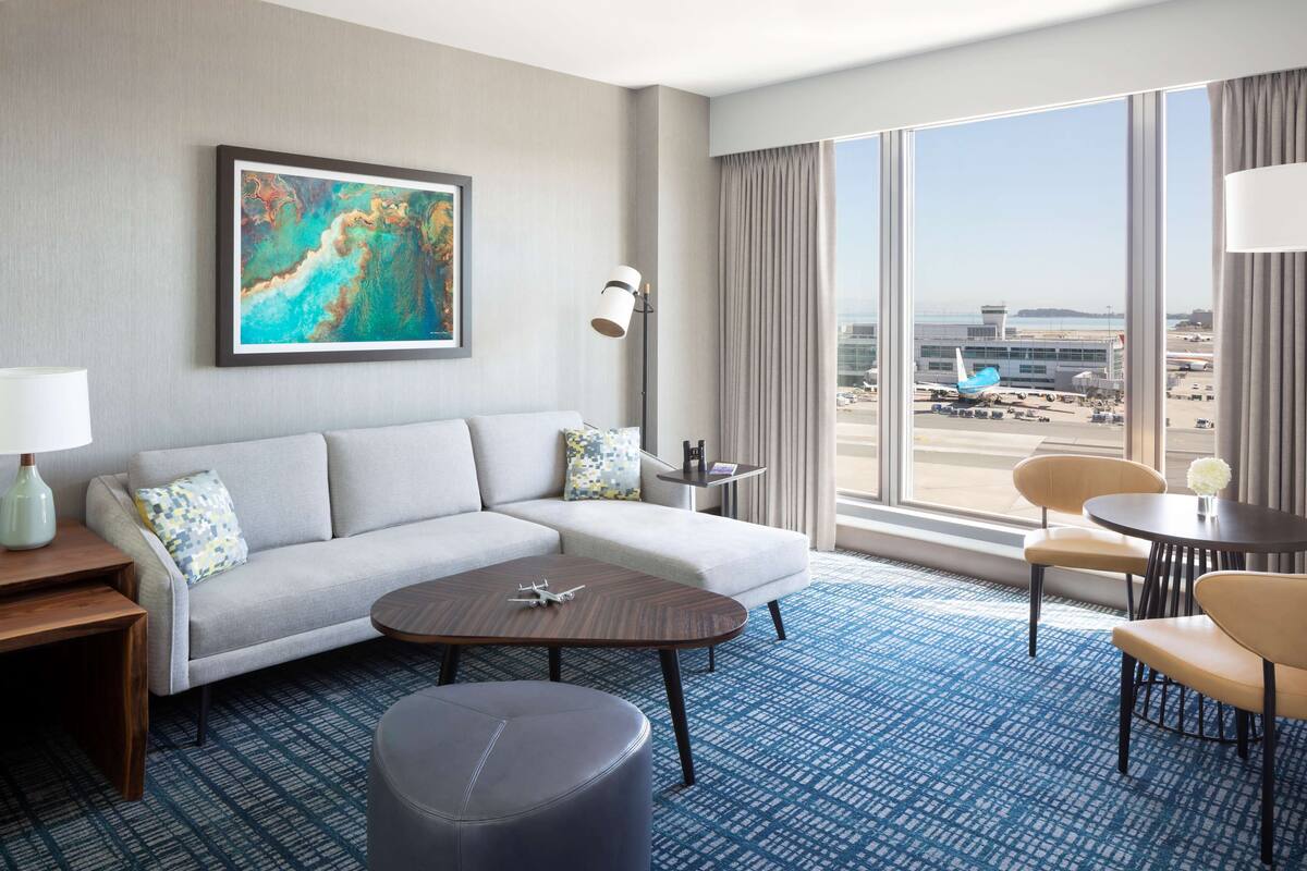 Habitación de hotel con un ventanal que ofrece vista a un aeropuerto, un sofá, una mesa de centro y un pequeño comedor.