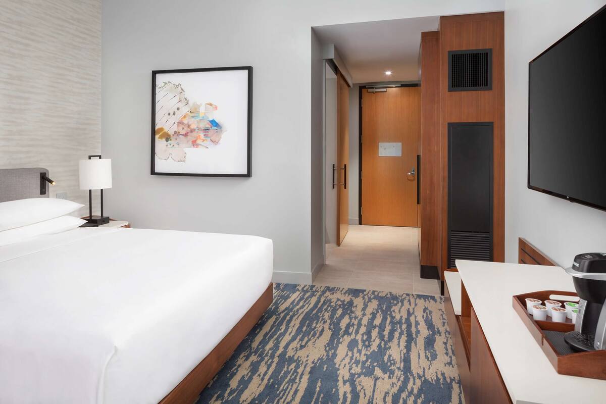 Una habitación de hotel con cama, escritorio, cafetera, televisor y un cuadro en la pared.