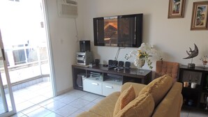 A living room with a sofa, a flat-screen TV mounted on the wall, a small table with speakers, and a decorative vase with flowers. - Praia das Dunas Apartamento Aconchegante 2 Quartos (Cabo Frio)