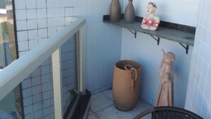 A small balcony with tiled walls, a wicker chair, a large terracotta pot, and a shelf with decorative items. - Praia das Dunas Apartamento Aconchegante 2 Quartos (Cabo Frio)