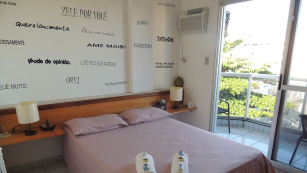 A hotel room with a bed, pillows, a nightstand with a lamp, a wall with motivational quotes, and a balcony with a view of greenery. - Praia das Dunas Apartamento Aconchegante 2 Quartos (Cabo Frio)