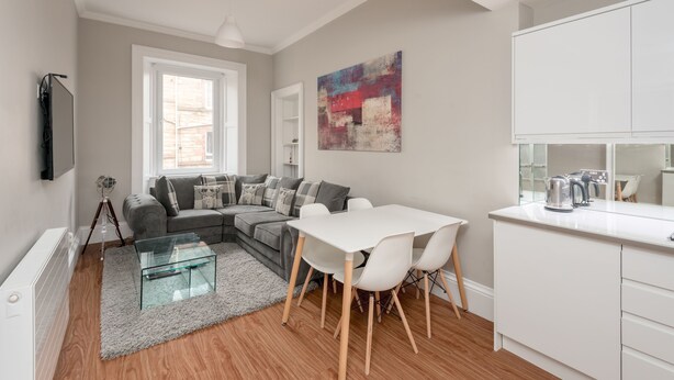 Una sala de estar moderna con un sofá gris, una mesa de centro de vidrio y una mesa de comedor blanca con sillas. Hay una zona de cocina con armarios blancos y una tetera sobre la encimera.