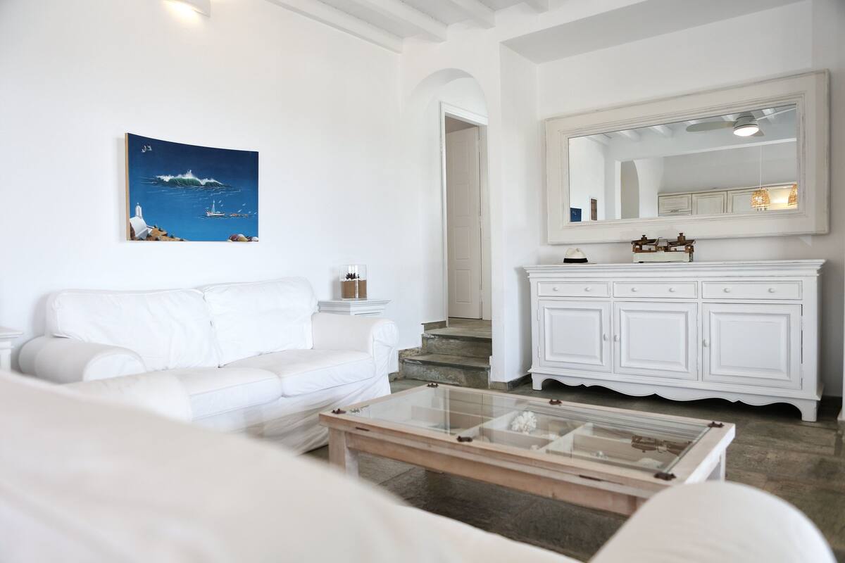 A white living room with a white sofa, a glass coffee table, a white cabinet, and a painting of a seascape.