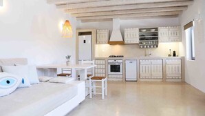 A compact, modern kitchen with white cabinetry, a dining area with a white table and chairs, and a cozy seating area with a sofa and cushions. - VLIA MAR Myconian Residences (Mykonos)
