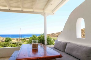 A wooden table with a glass and a plant, a white wall with a window, and a view of the sea and hills. - VLIA MAR Myconian Residences (Mykonos)