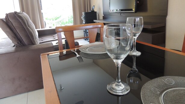 A dining area with a glass table, two wine glasses, and a television mounted on the wall. - Casa Cabo Frio Aconchegante 5min Praias (Cabo Frio)