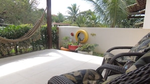 A wicker chair with a camouflage cushion, a hammock, and potted plants on a balcony with a white floor. - Casa Cabo Frio Aconchegante 5min Praias (Cabo Frio)