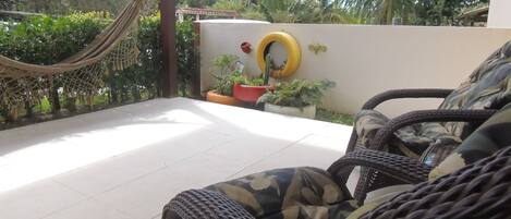 A wicker chair with a camouflage cushion, a hammock, and potted plants on a balcony with a white floor.