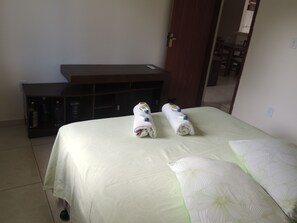 A hotel room with a bed, a small wooden side table with bottles, and a door leading to a dining area.