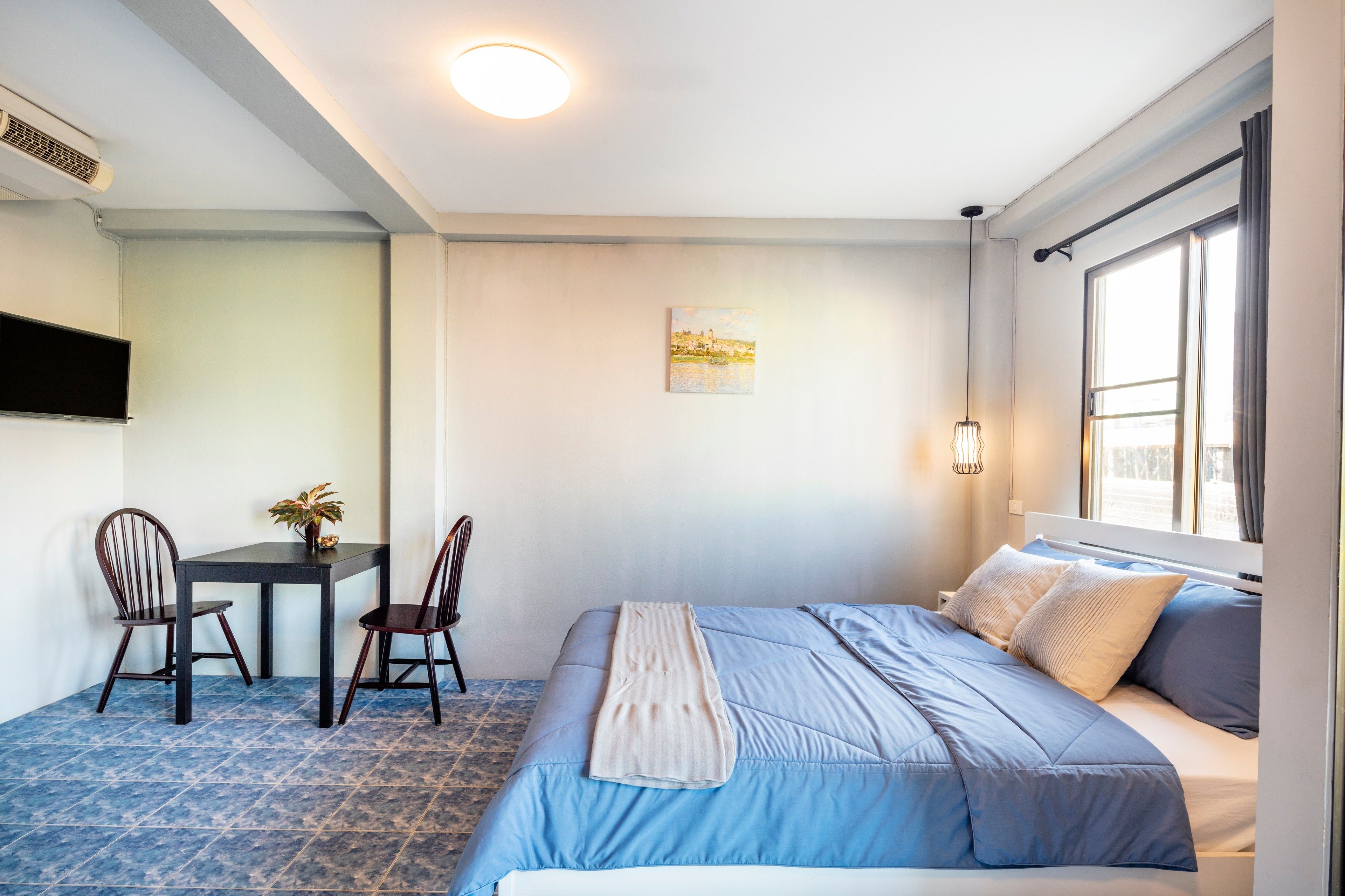Une chambre à coucher avec un lit, un bureau, deux chaises, une plante en pot, un tableau et une fenêtre avec des rideaux.