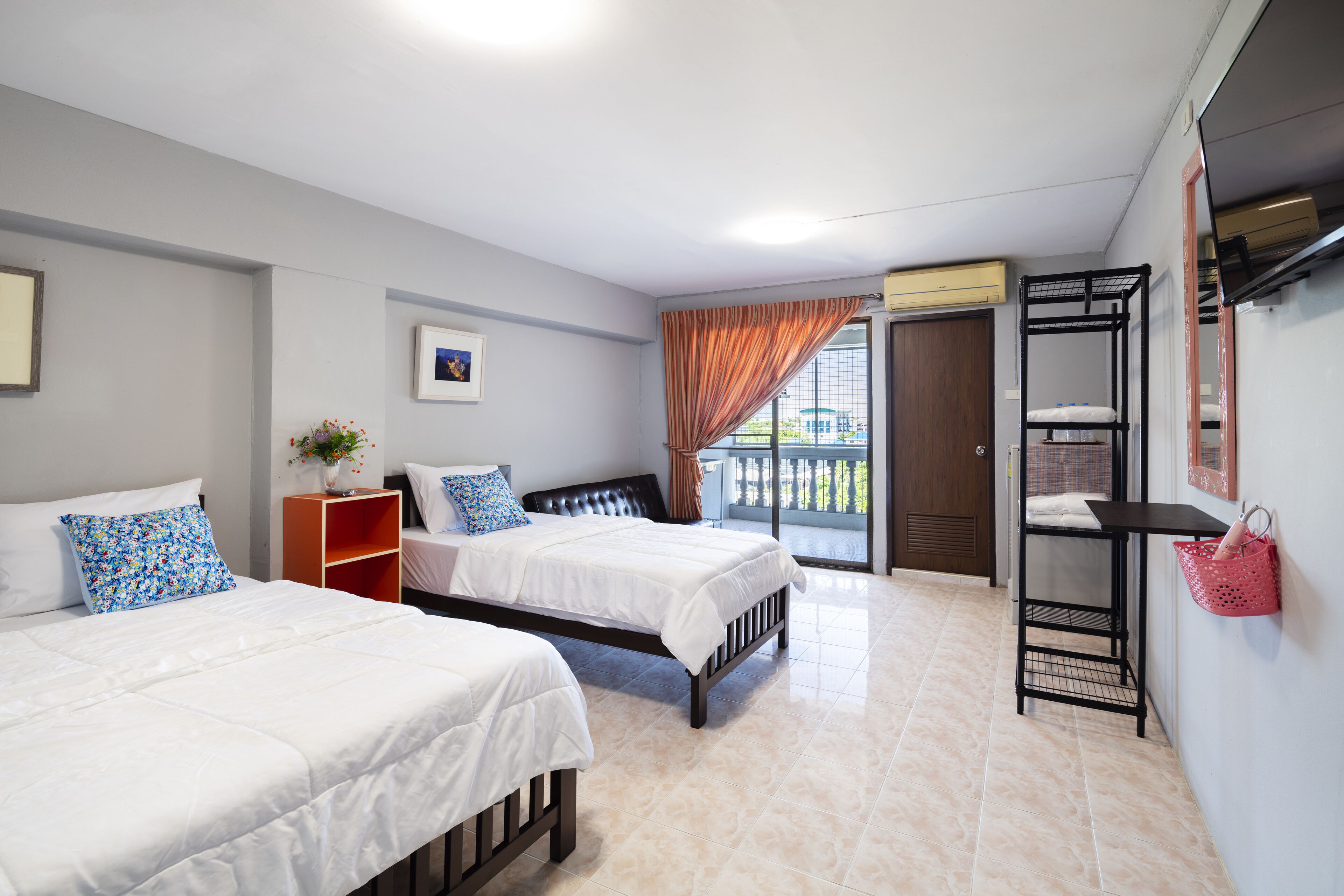 A hotel room with two beds, a small table with a vase, a shelf with towels, and a balcony with a view.