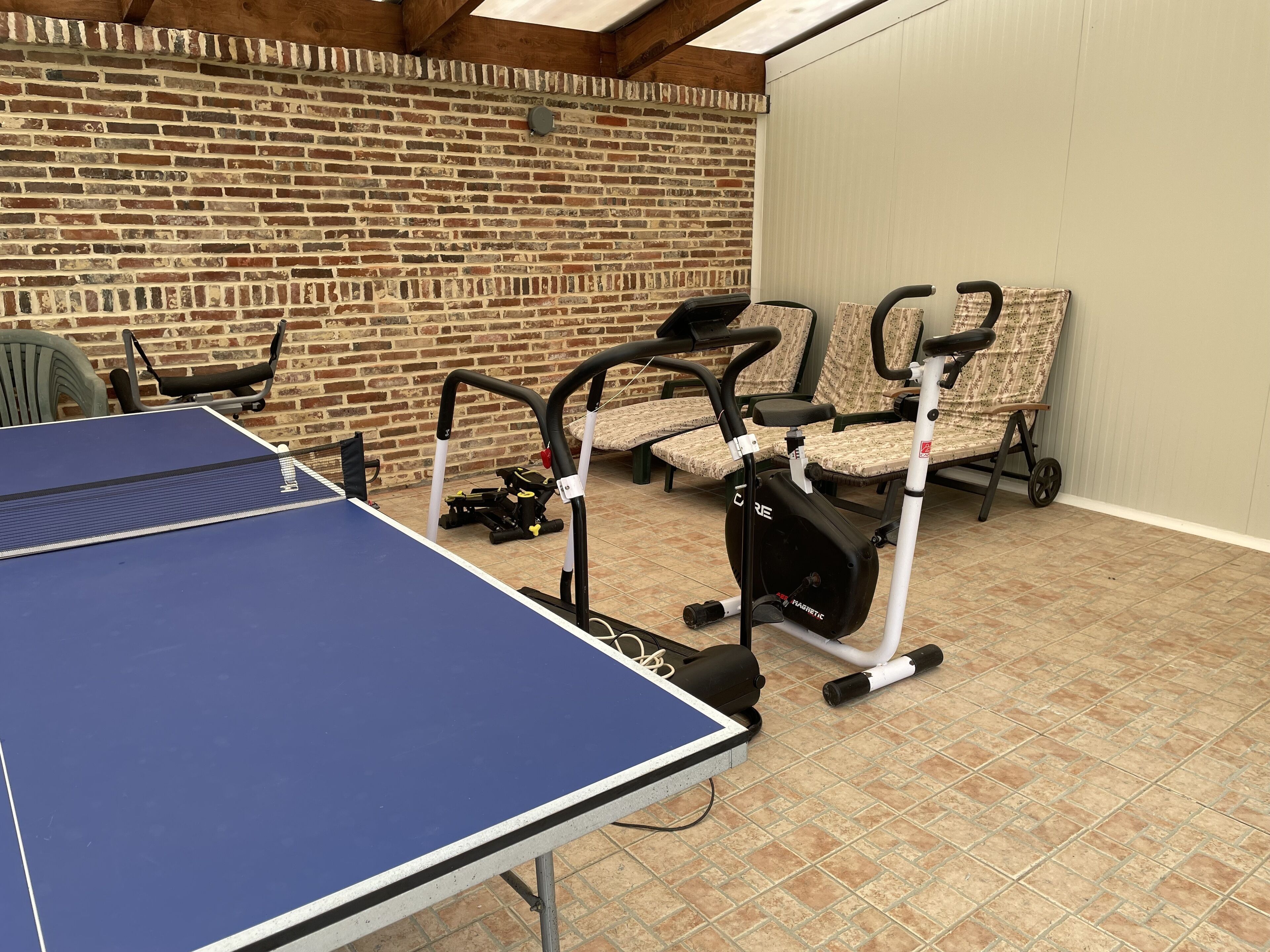 A room with a ping pong table, exercise equipment, and a brick wall.
