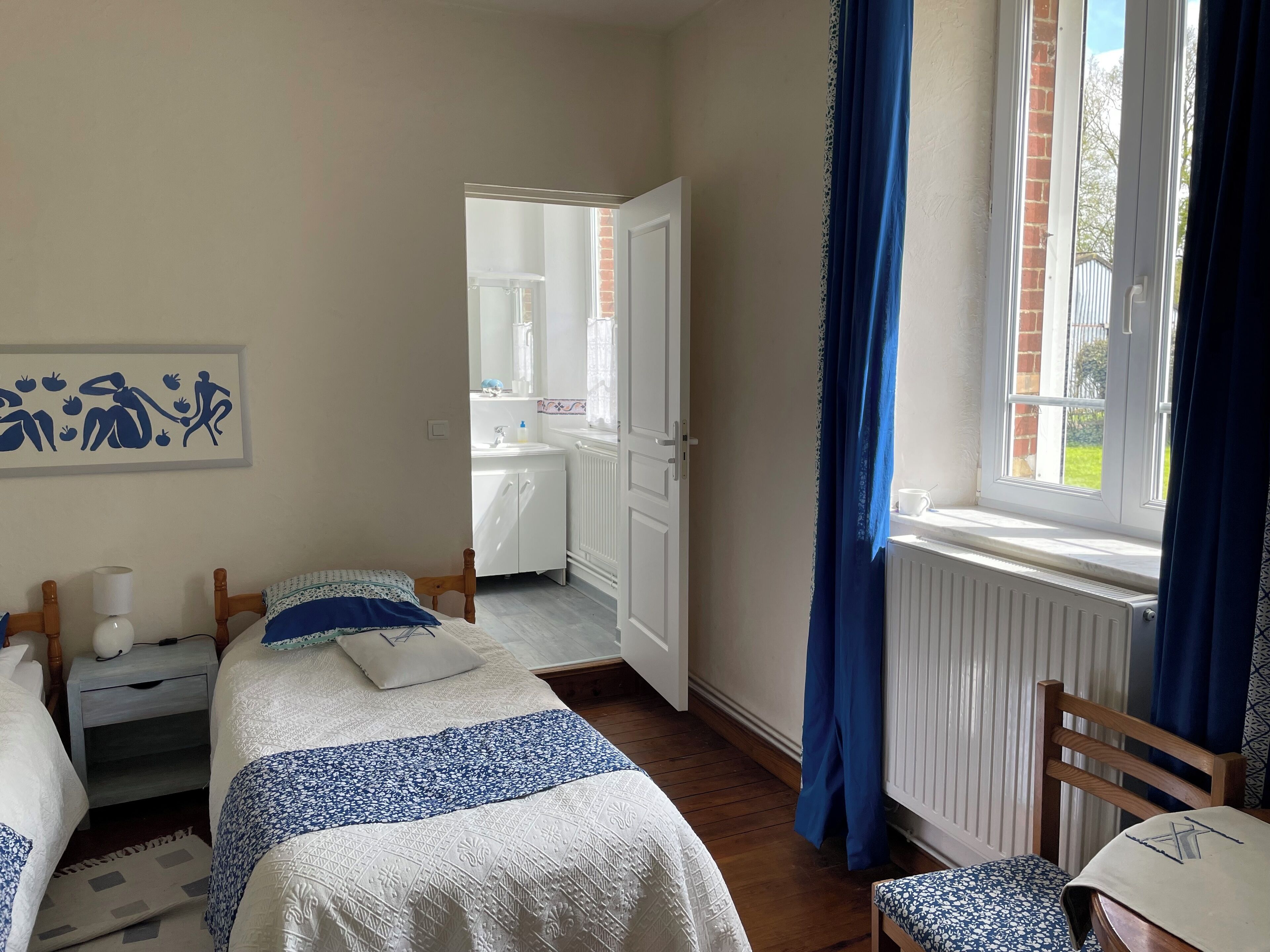 A bedroom with two beds, a bedside table with a lamp, a window with blue curtains, and a view of a bathroom.