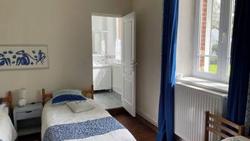 A bedroom with two beds, a bedside table with a lamp, a window with blue curtains, and a view of a bathroom.