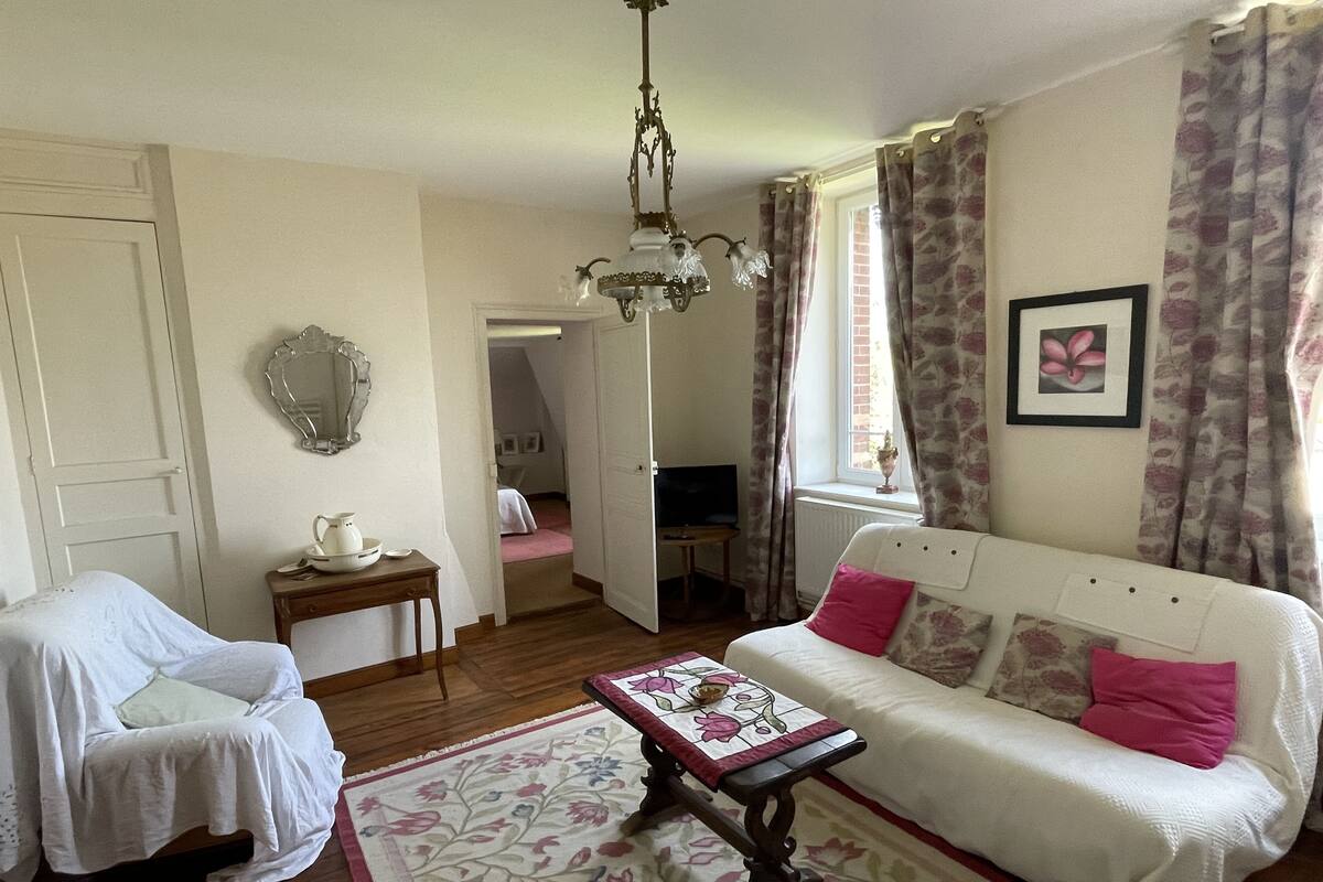 A cozy room with a white sofa, a small table with a floral table runner, a mirror on the wall, and a chandelier.