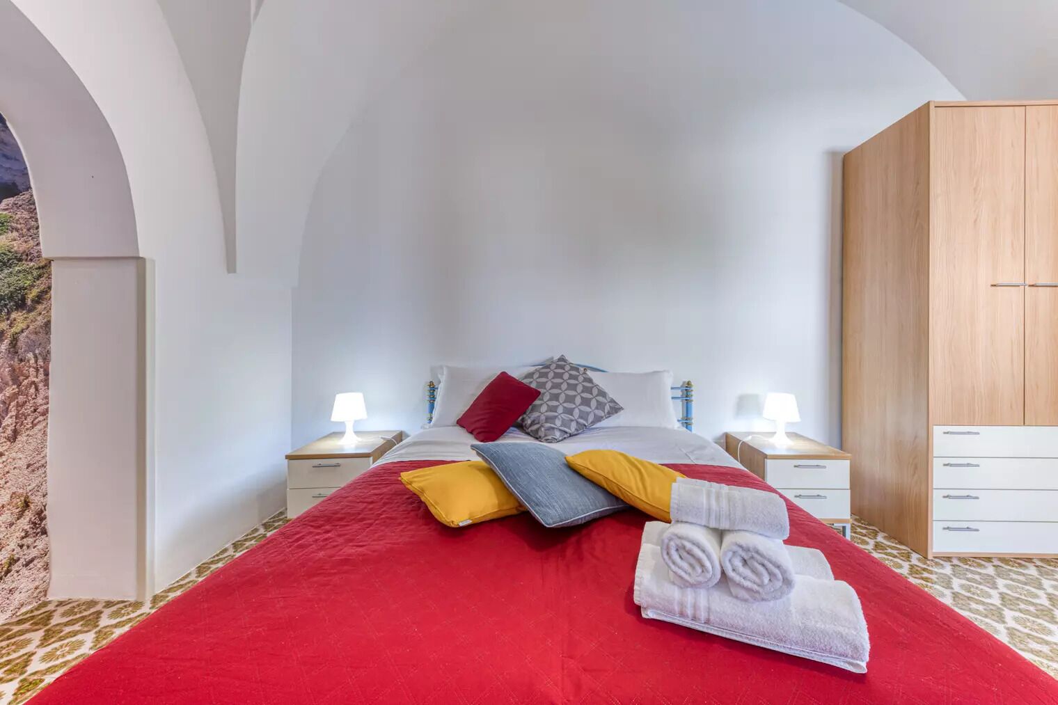 A bedroom with a large bed, two bedside tables with lamps, a wooden wardrobe, and a view of the outdoors through an arched window.