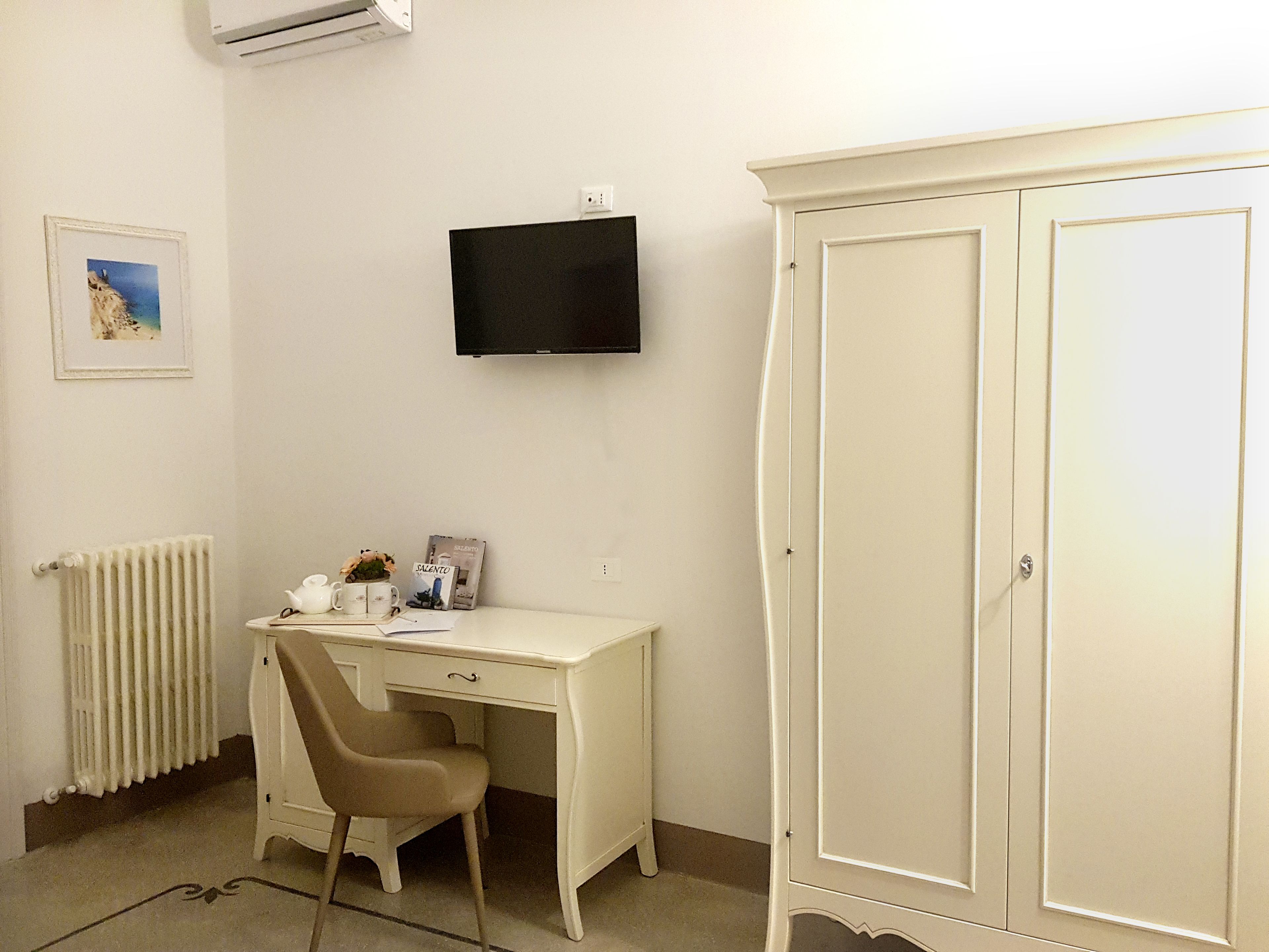 Ein Zimmer mit einem weißen Schreibtisch, einem Stuhl, einem Heizkörper, einem an der Wand montierten Fernseher und einem großen Kleiderschrank.