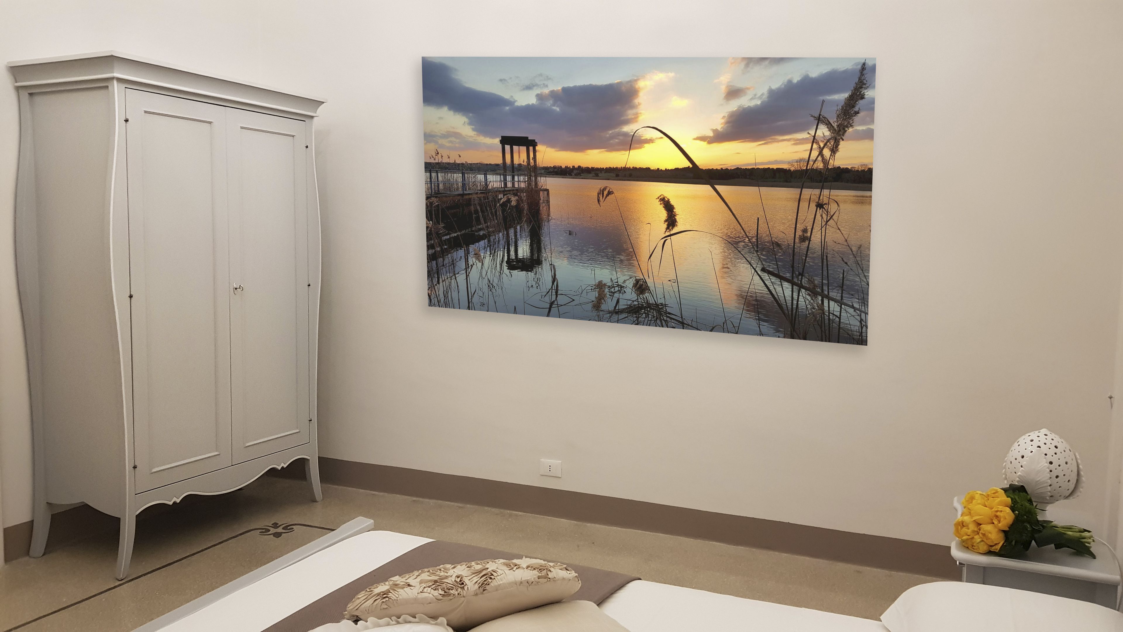 Een kamer met een witte kast, een bed met een bloemenkussen, en een schilderij van een zonsondergang boven een meer.