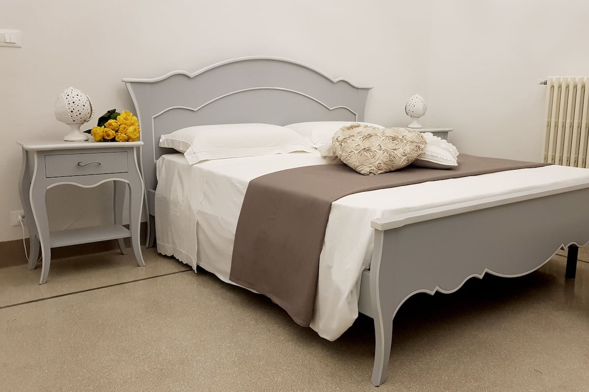 Una cama bien hecha con estructura gris, ropa de cama blanca y una manta marrón. Hay una mesita de noche pequeña con una lámpara y un jarrón con flores amarillas. Se ve un radiador en la pared, y un reposapiés beige está colocado junto a la cama.