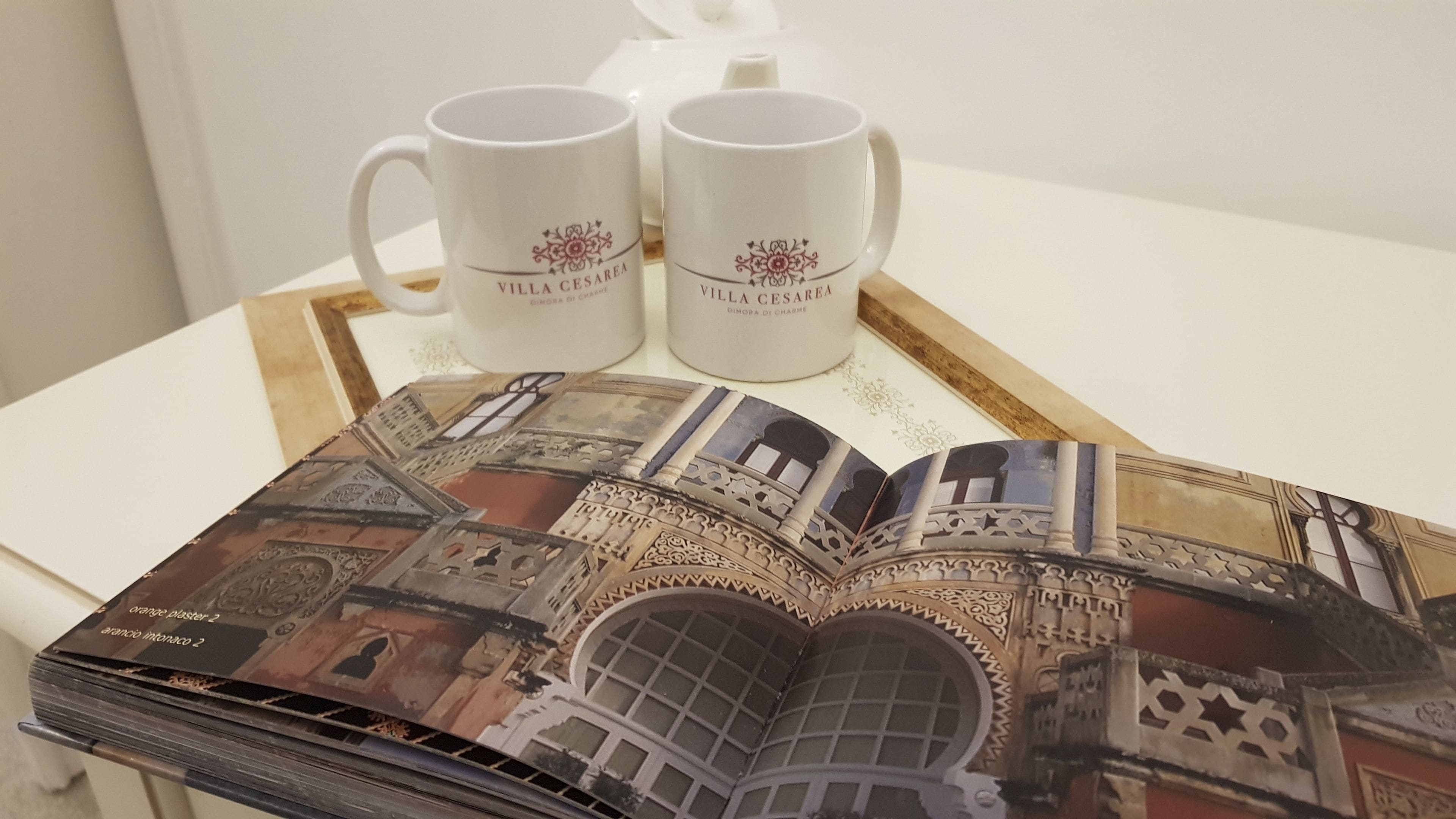 A book with architectural illustrations, two white mugs with "Villa Cesarea" text, and a tray on a table.