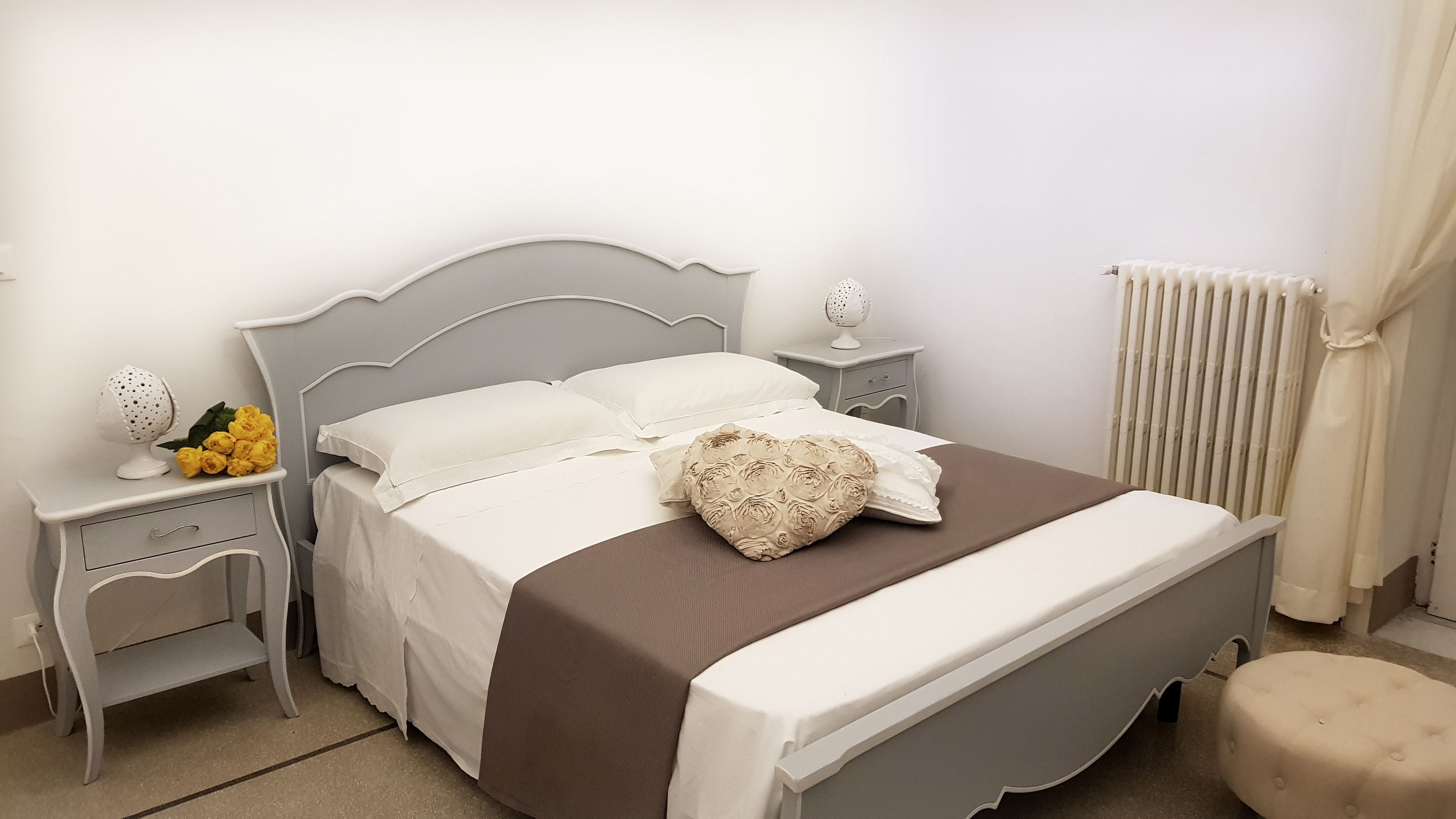 A neatly made bed with a brown bedspread and white pillows, a bedside table with a lamp and a decorative item, and a radiator on the wall.