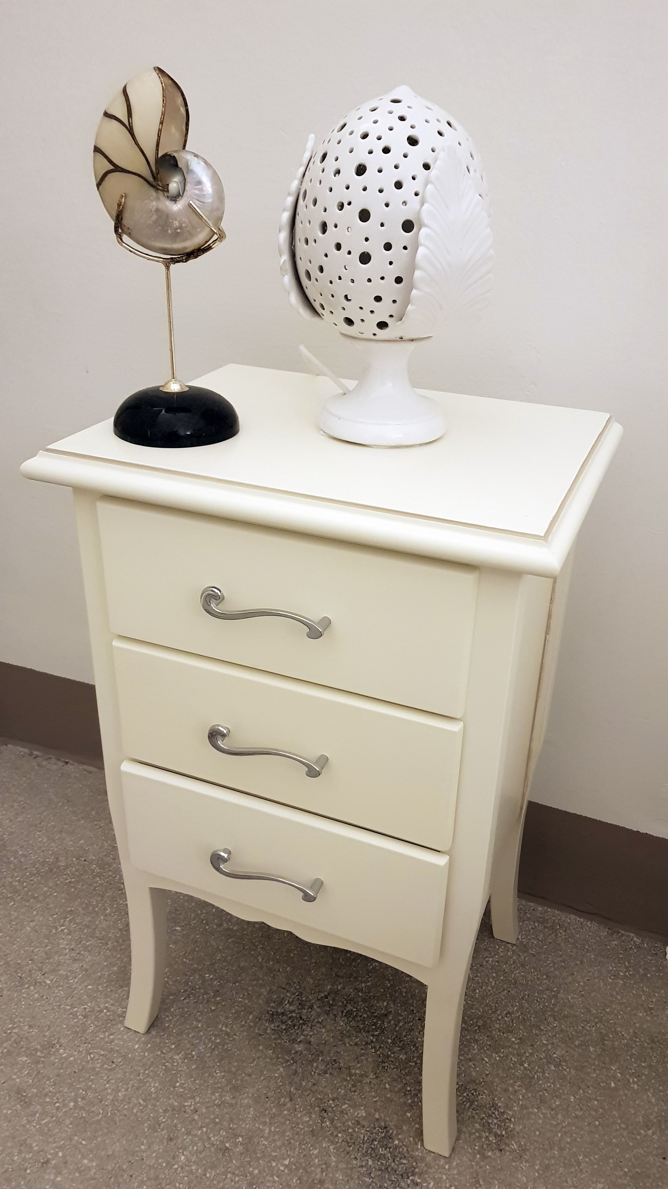 Una mesita de noche blanca con tres cajones y dos objetos decorativos encima.