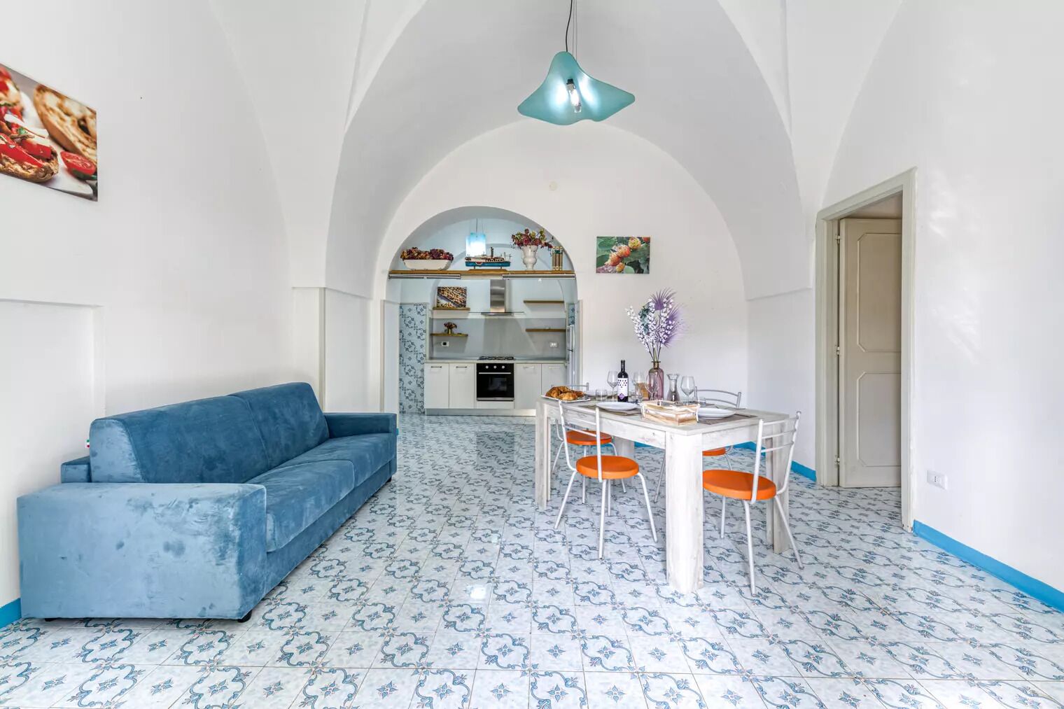 Una sala de estar moderna con un sofá azul, una mesa de comedor con sillas naranjas y una cocina al fondo.