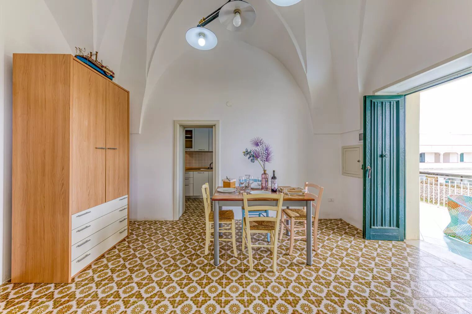 A dining area with a table set for two, a wooden wardrobe, and a kitchen in the background.
