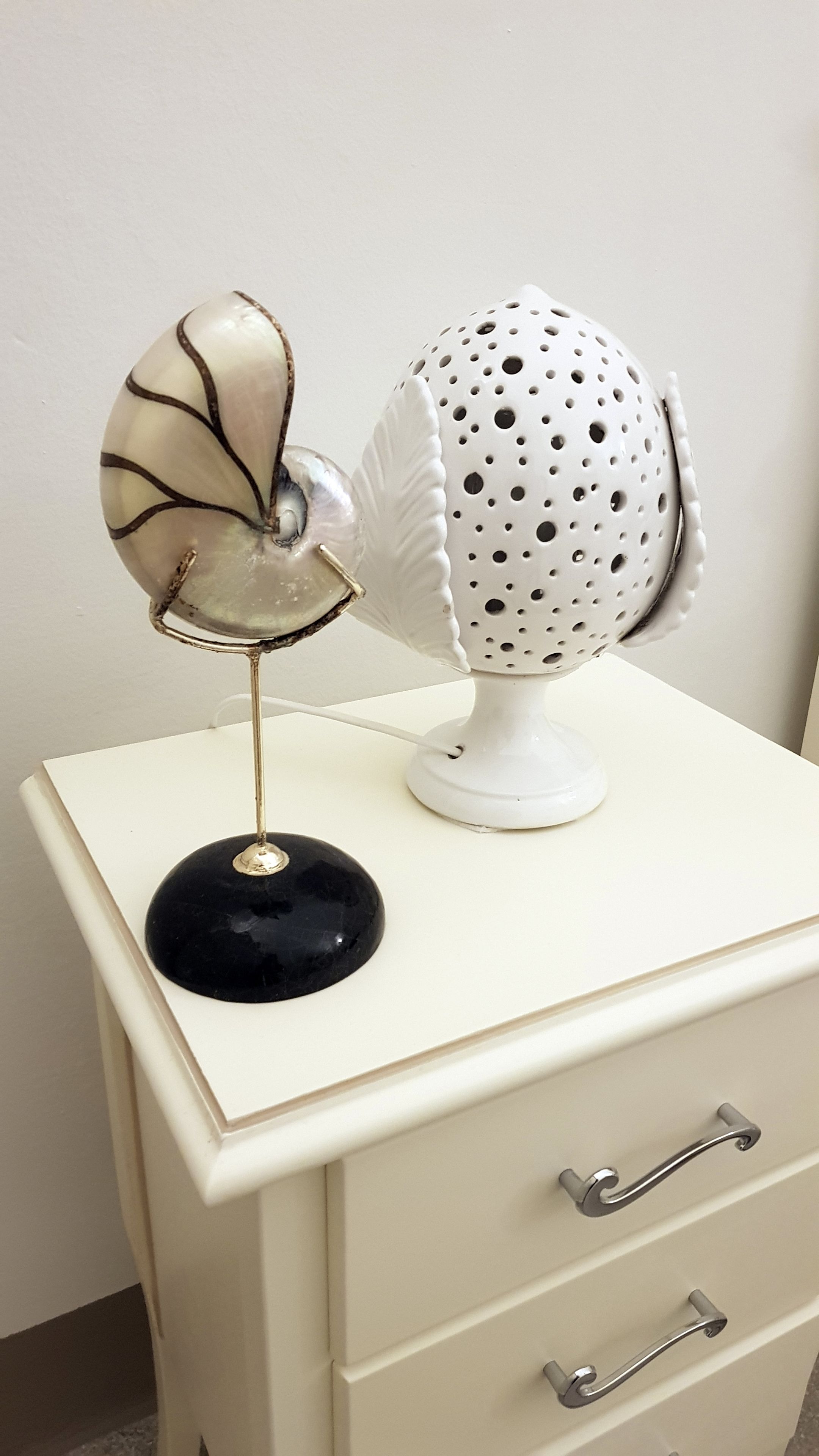 Une lampe de style vintage, dotée d’un pied métallique et d’un abat-jour décoratif, posée sur une commode blanche avec des tiroirs et un socle noir.