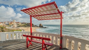 A red bench under a shelter by the seaside, overlooking a rocky coastline and buildings. - HI Santa Cruz - Pousada de Juventude - Hostel (Torres Vedras)