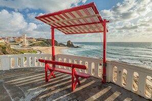 Un banc rouge, placé sous un abri en bord de mer, d’où l’on peut admirer une côte rocheuse et des bâtiments.