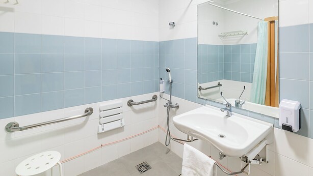 Un baño con lavamanos blanco, un espejo, un cabezal de ducha de mano y un perchero para toallas.